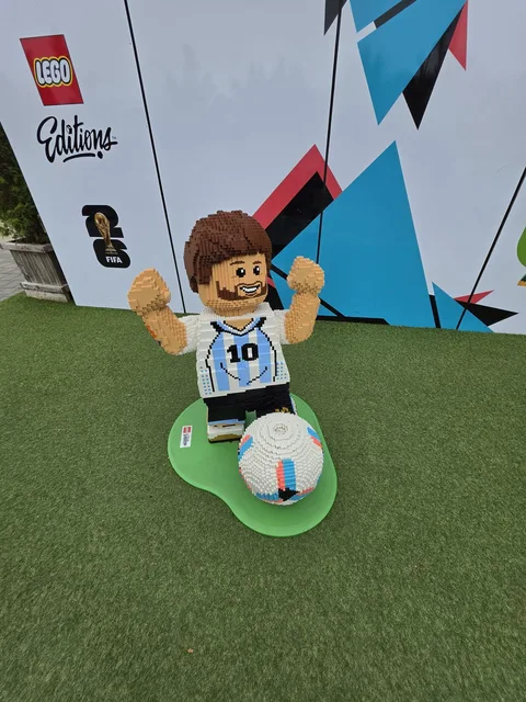 A brick-built football player figure in a light-blue and white striped number 10 shirt, arms raised, with a ball at his feet, in front of a LEGO Editions backdrop