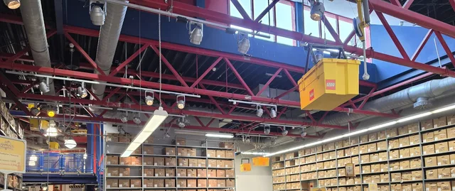 Inside the LEGO Fabrik Shop: rows of red and blue bins of loose LEGO elements beneath a red steel roof, shelves of boxes along the walls, and a giant yellow LEGO storage box hanging from the ceiling