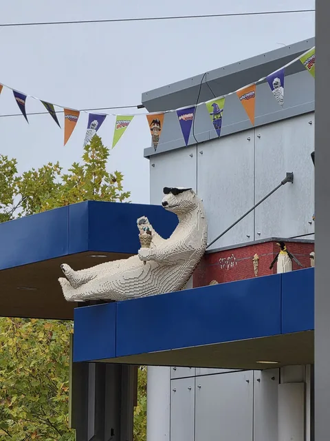 A large brick-built polar bear in sunglasses lying back on a blue roof edge, holding an ice-cream cone, with Halloween bunting strung above him and a brick penguin beside him