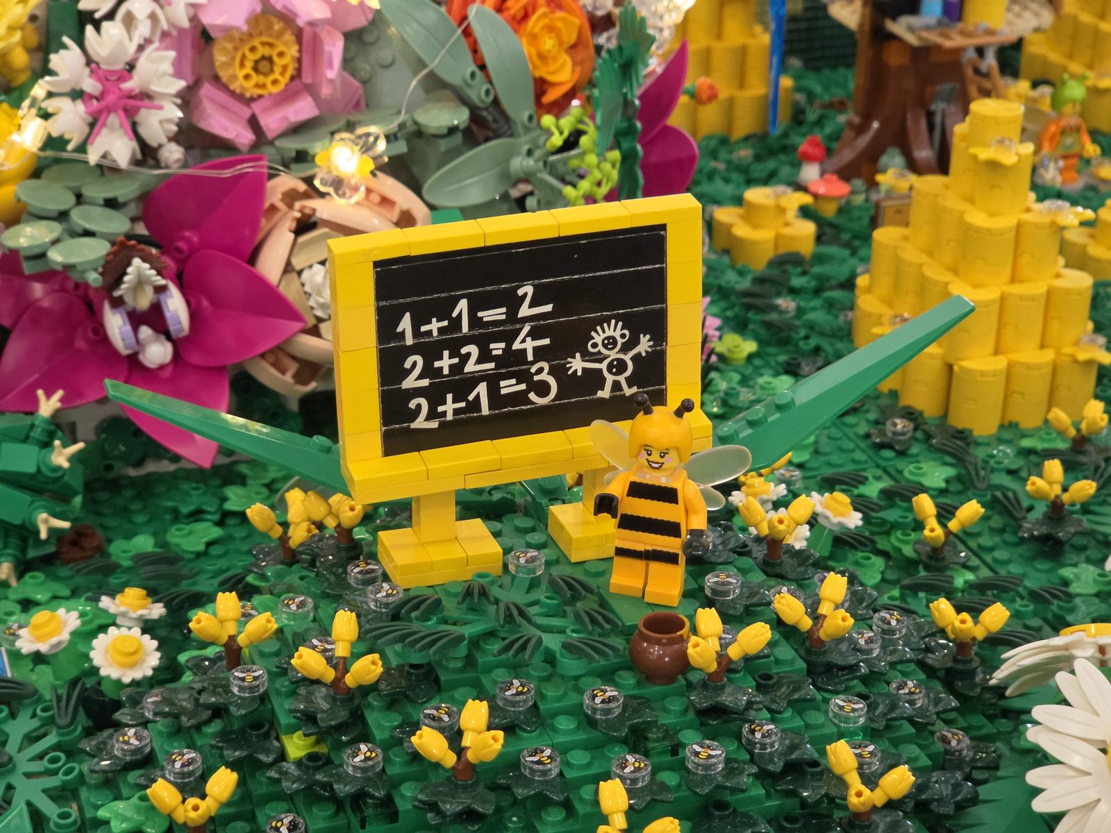A small LEGO blackboard in a flower meadow, carrying three sums and a chalk stick figure; in front of it a minifigure in a bee costume