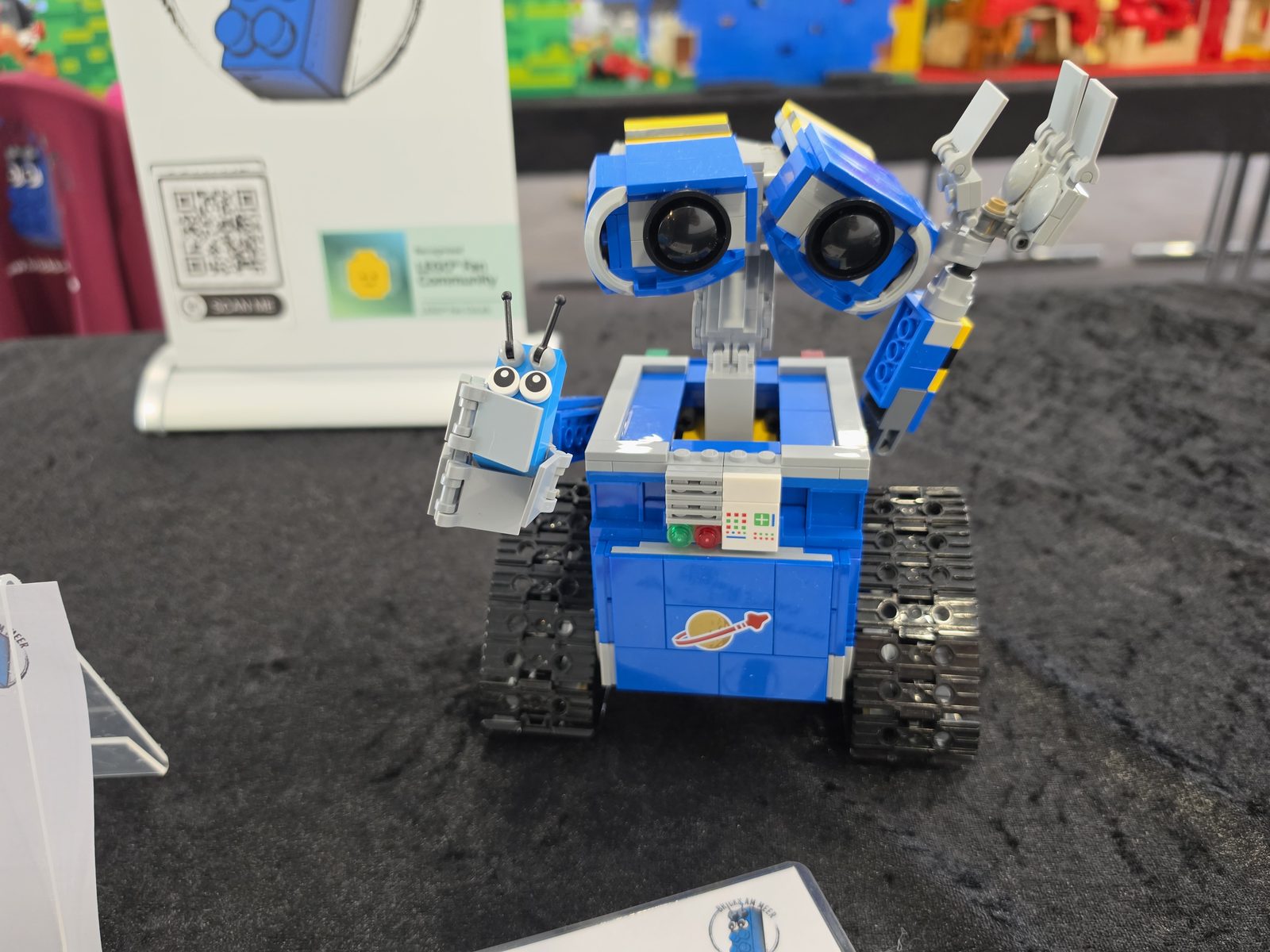 A small blue robot built from LEGO bricks with large eyes, beside the Bricks am Meer mascot